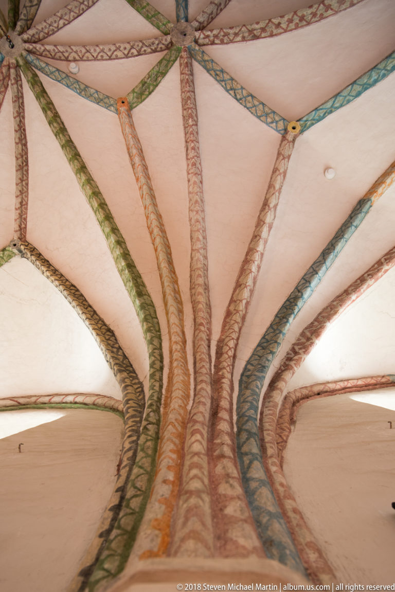 Close upward view of painted red, green, blue, and ochre vault ribs fanning across a pale ceiling.