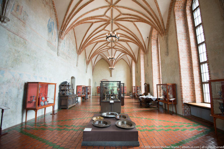 Long vaulted castle gallery with display cases, tall windows, red-and-green tile floor, and red-brick ceiling ribs.