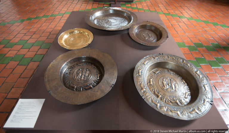 Five embossed metal plates of different sizes and tones arranged on a low display plinth over a red-and-green tiled floor.