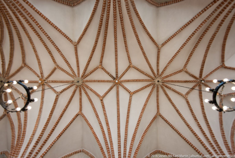 White vaulted ceiling crossed by red-brick ribs forming repeated star patterns, with dark circular chandeliers below.