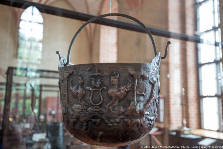 Dark patinated metal cauldron with arched handle and embossed heraldic or figural decoration displayed in a museum case.