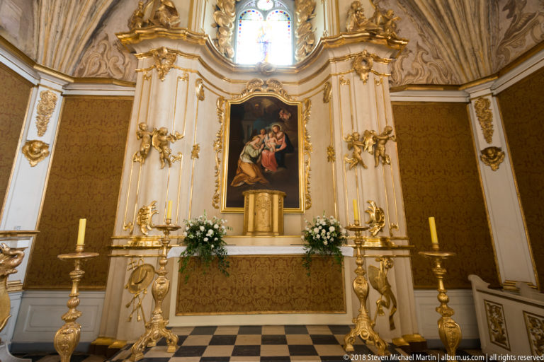 Baroque-style altar with central religious painting, gilded ornament, flowers, and tall candlesticks beneath a window.
