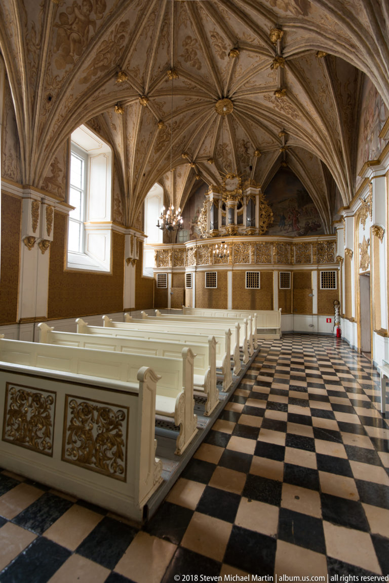 Long vaulted chapel-like room with white benches, black-and-white checkerboard floor, chandeliers, and gilded decoration.