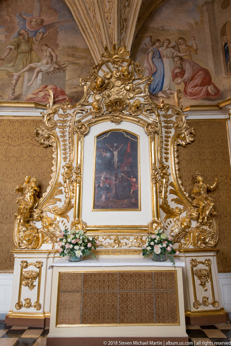Small religious painting centered within an elaborate white-and-gold Rococo wall surround with sculpted figures and painted decoration.