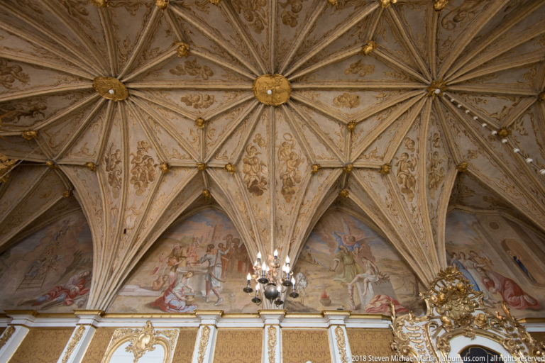 Decorated rib vault with gilded bosses, painted ornament, and mural scenes above an ornate interior wall.