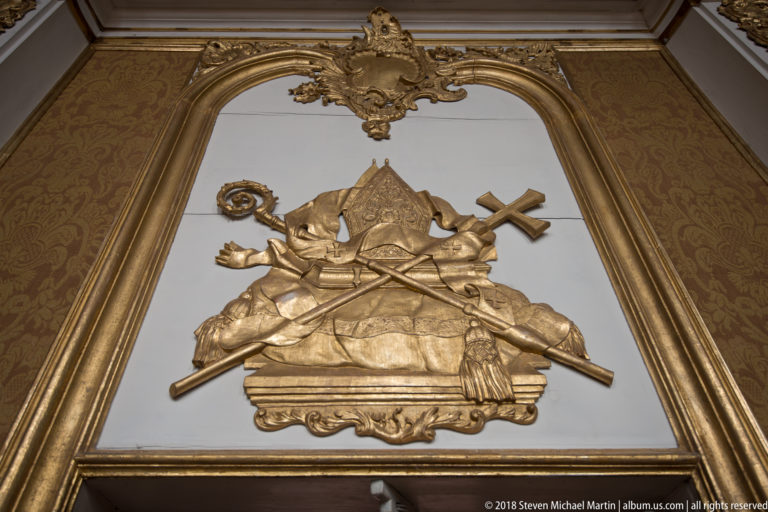 Gilded ecclesiastical emblems with crossed staffs and a papal-style headdress fill an ornate framed wall panel.