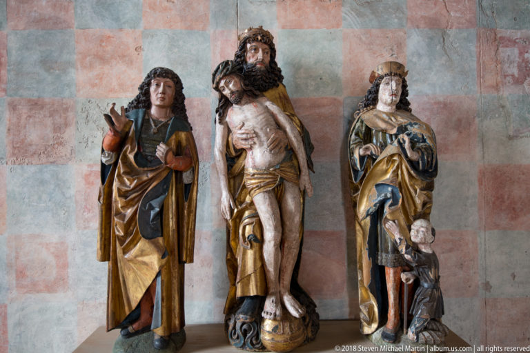Three polychrome wooden religious sculptures stand side by side against a pale red-and-gray checker-patterned wall.