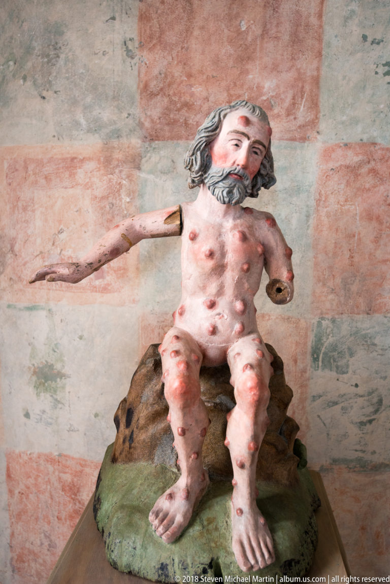 Polychrome sculpture of a bearded nearly nude man seated on a rock with red sores and damaged forearms