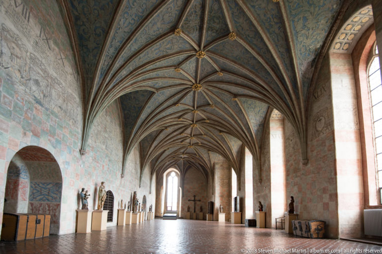 Long Gothic hall viewed toward a distant doorway, with ribbed vaults, tall windows, painted walls, and sculptures