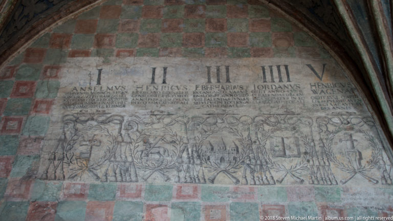 Faded wall painting with Roman numerals, Latin names, and rows of small figures on a checker-patterned wall