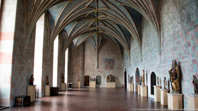Long vaulted hall with tall windows, painted walls, statues on plinths, and patterned floor inside Lidzbark Warmiński Castle