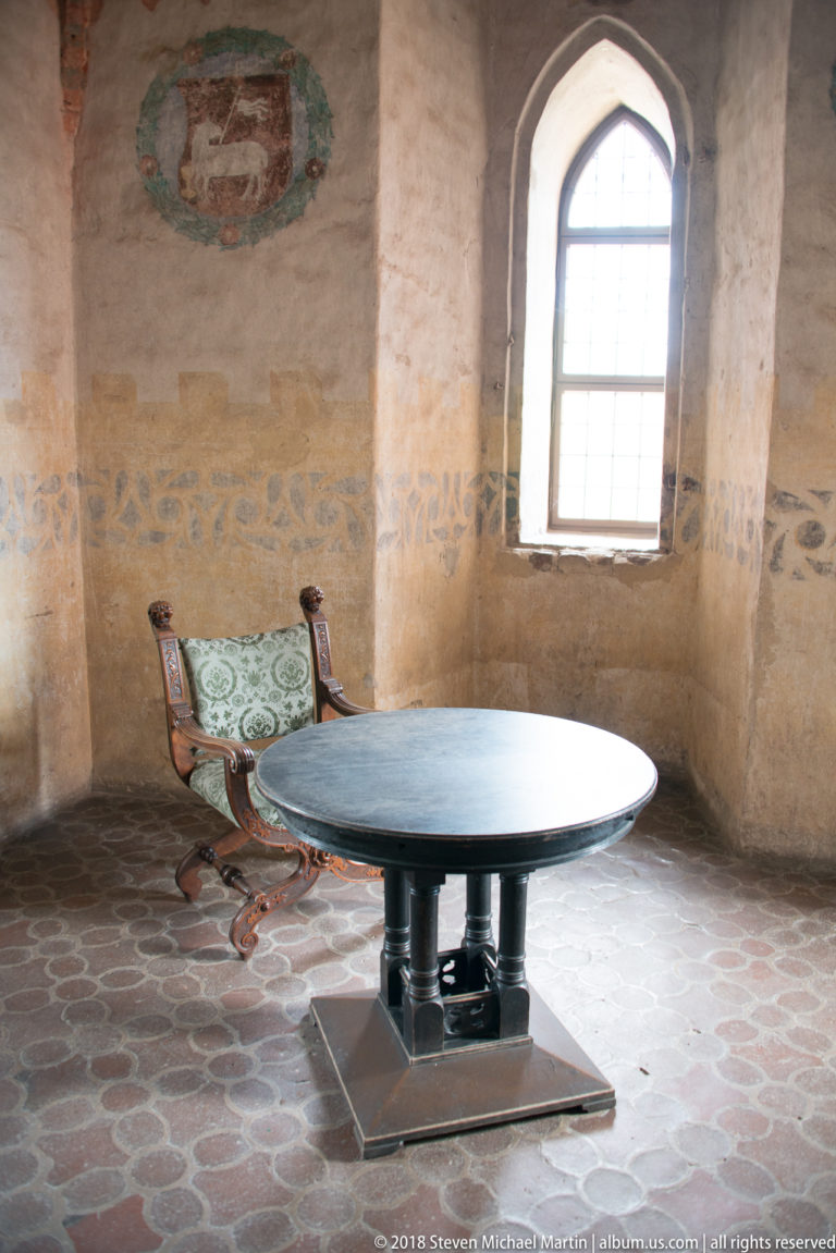 Round table and carved chairs in a pale castle room with pointed-arch window, tiled floor, and faded wall painting