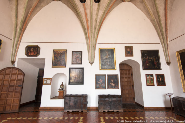 Historic paintings and heavy wooden chests line a white wall beneath Gothic vault ribs inside Lidzbark Warmiński Castle