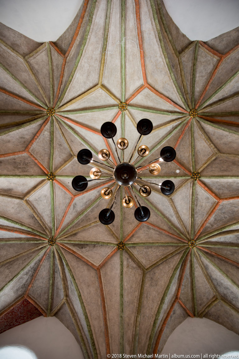 Upward view of a Gothic star vault with red and green ribs, small bosses, and a modern circular ceiling light