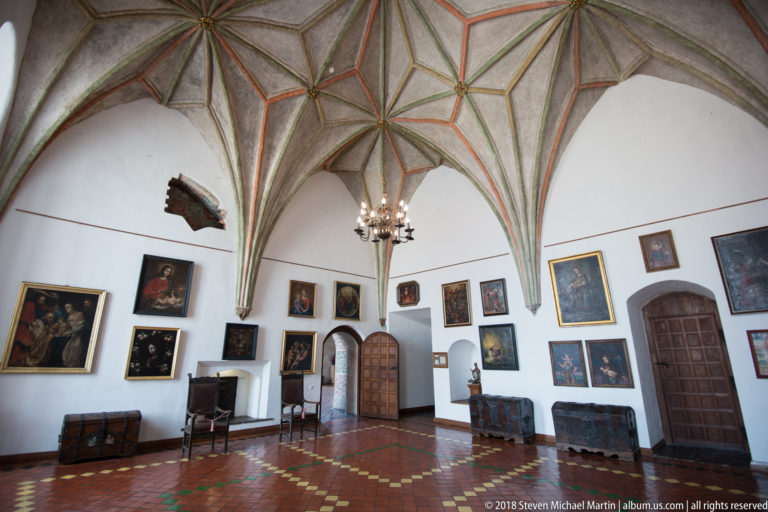 White-walled Gothic hall with star vault, historic paintings, wooden doors, chests, and a hanging chandelier