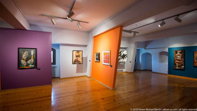 Contemporary art gallery with purple, orange, and blue walls, framed paintings, track lights, and polished wooden floor