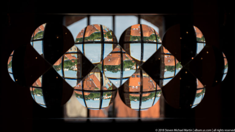 Dark circular forms overlap a window, fragmenting bright red roofs outside into repeated geometric shapes