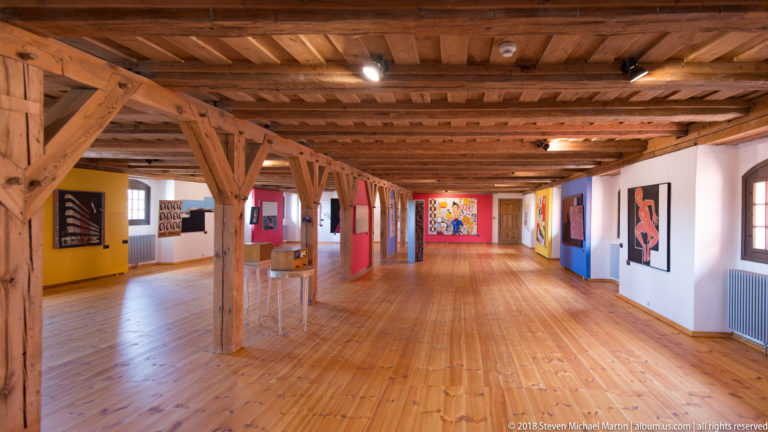Long gallery with wooden floor and beams, colorful partitions, and contemporary paintings inside Reszel Castle