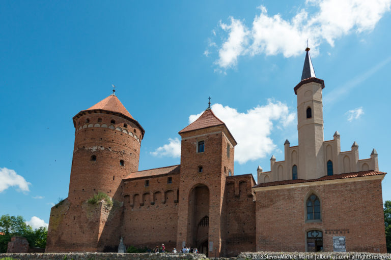 Reszel Castle exterior with massive round brick tower, square gate tower, and white gabled wing with slender spire