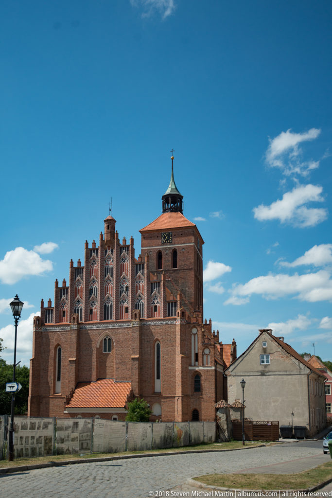 Broad side view of Reszel’s red-brick Gothic church with tower, stepped gables, buttresses, and tiled roof