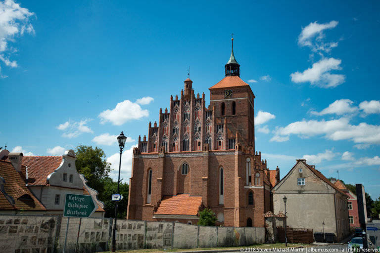 Brick Gothic church in Reszel behind a road sign, stone wall, and neighboring houses under blue sky