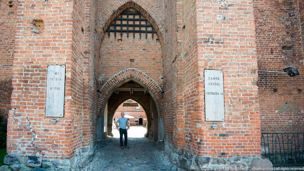 Pointed-arch brick gateway with a person walking through into a courtyard beyond at Reszel Castle