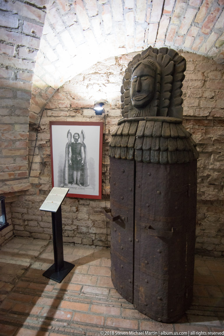 Large dark wooden barrel-like object covered with studs and topped by a carved head in a brick-vaulted exhibit room