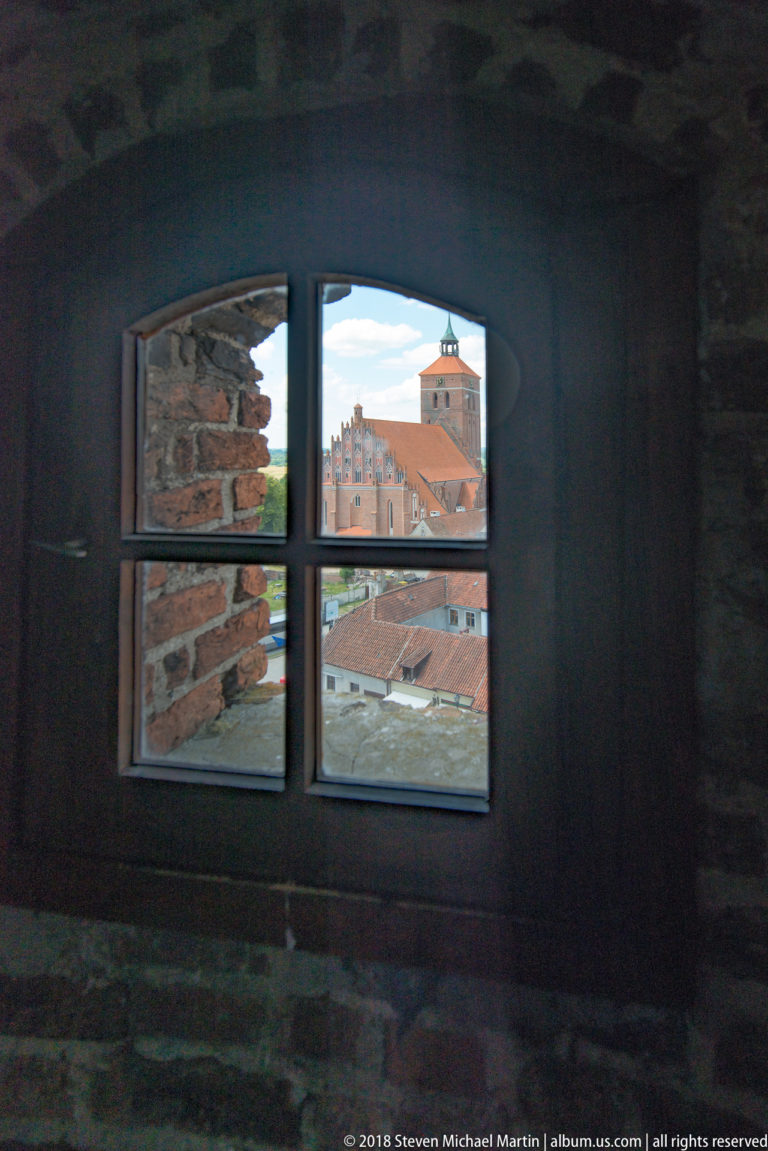 Bright church tower and red roofs of Reszel seen through a small multi-pane window in a dark castle interior