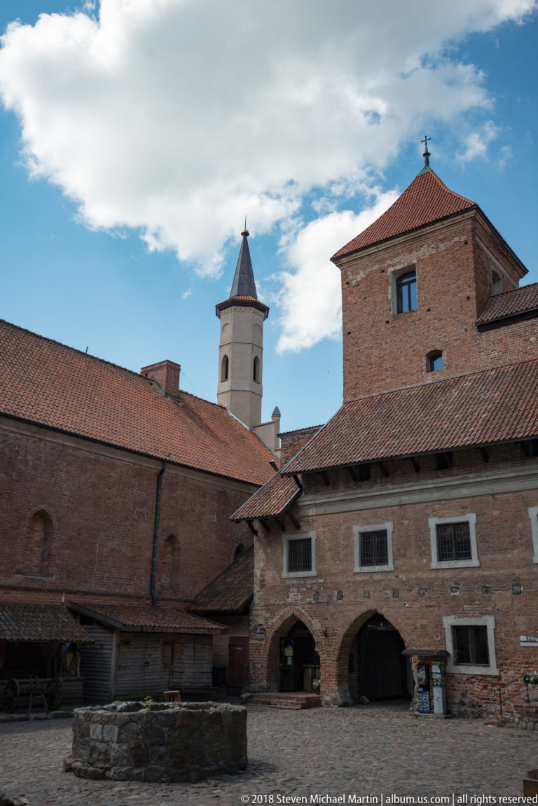 Brick courtyard ranges with arched openings and tiled roofs beneath a slender white spire at Reszel Castle
