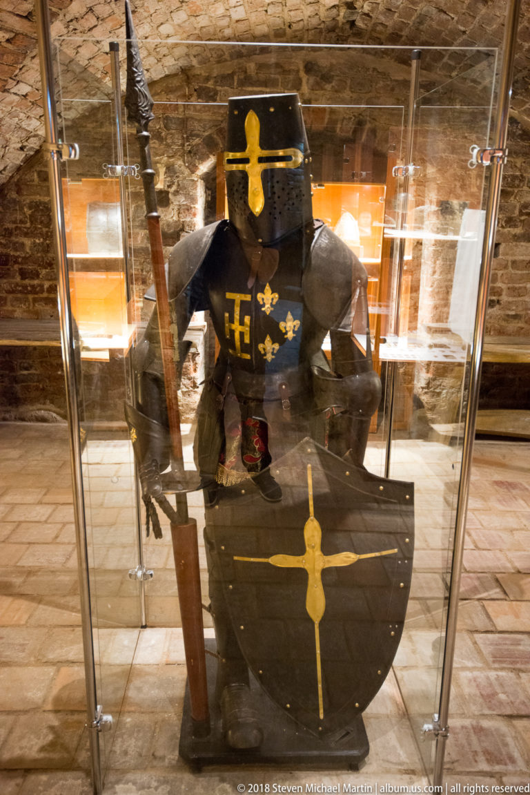 Black armor with gold decoration displayed behind a large black shield and upright weapons in a glass museum case