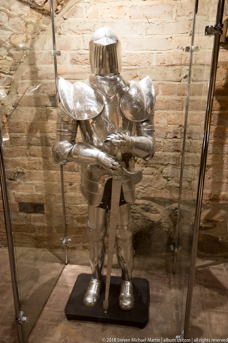 Full suit of polished plate armor displayed upright in a glass case against an exposed brick wall