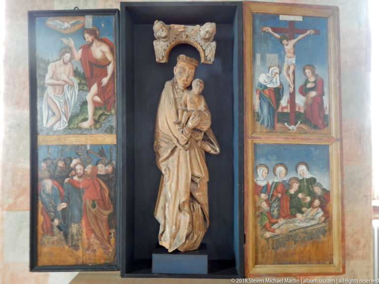 Carved Madonna and Child in a central niche flanked by painted religious panels with Passion scenes and saints.
