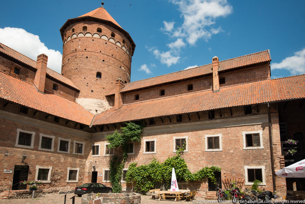 Courtyard of Reszel Castle with red-brick walls, orange tile roofs, timber galleries, ivy, and a large round tower