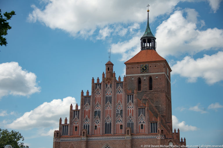 Close view of Reszel’s brick Gothic church tower, clock, stepped pinnacles, pointed windows, and copper-toned spire