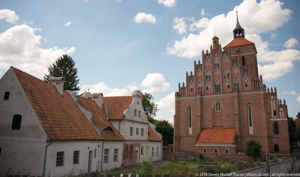 Brick Gothic church with ornate stepped gable and square tower beside low white buildings with red tile roofs in Reszel