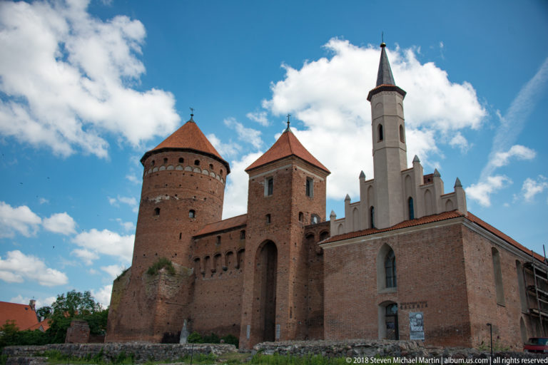 Red-brick Reszel Castle with a round tower, gate tower, tiled roofs, and a white gabled wing under blue sky