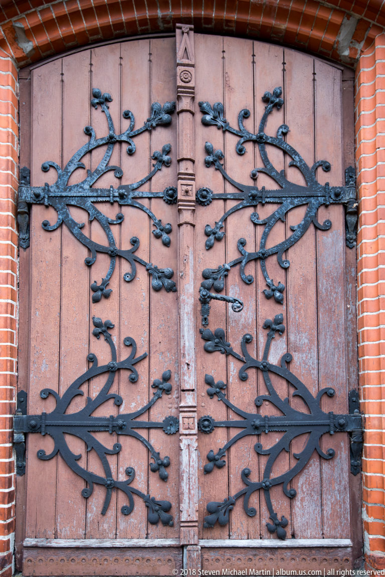 Wooden double church doors with symmetrical black iron strap hinges branching into decorative leaf-like forms.