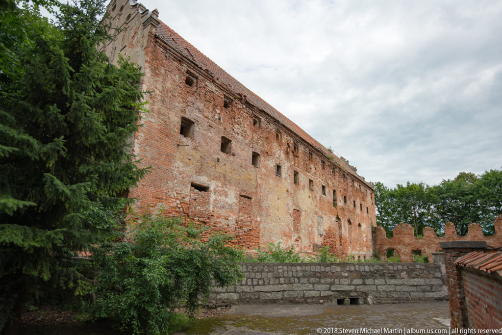 Long weathered brick range of the Warmian Chapter Castle in Pieniężno seen above a stone wall and trees.