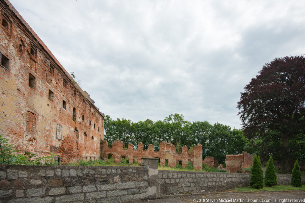 Stone boundary wall in front of the weathered brick range and broken crenellations of Pieniężno’s Warmian Chapter Castle.
