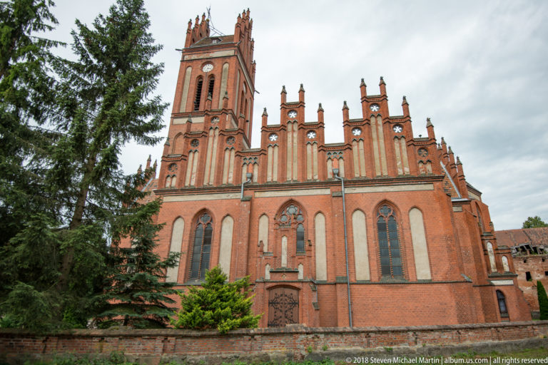 Red-brick Church of Saints Peter and Paul in Pieniężno with tall west tower, pointed windows, buttresses, and stepped gables.