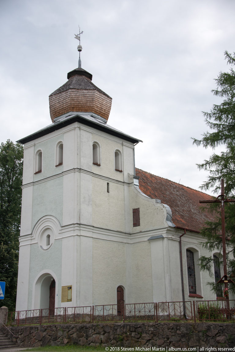 White masonry church with a broad square tower, small cross, arched openings, and a long red-tiled nave.