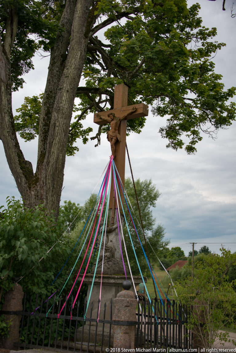 Wooden roadside crucifix beside a large tree, decorated with long multicolored ribbons tied toward a low metal fence.
