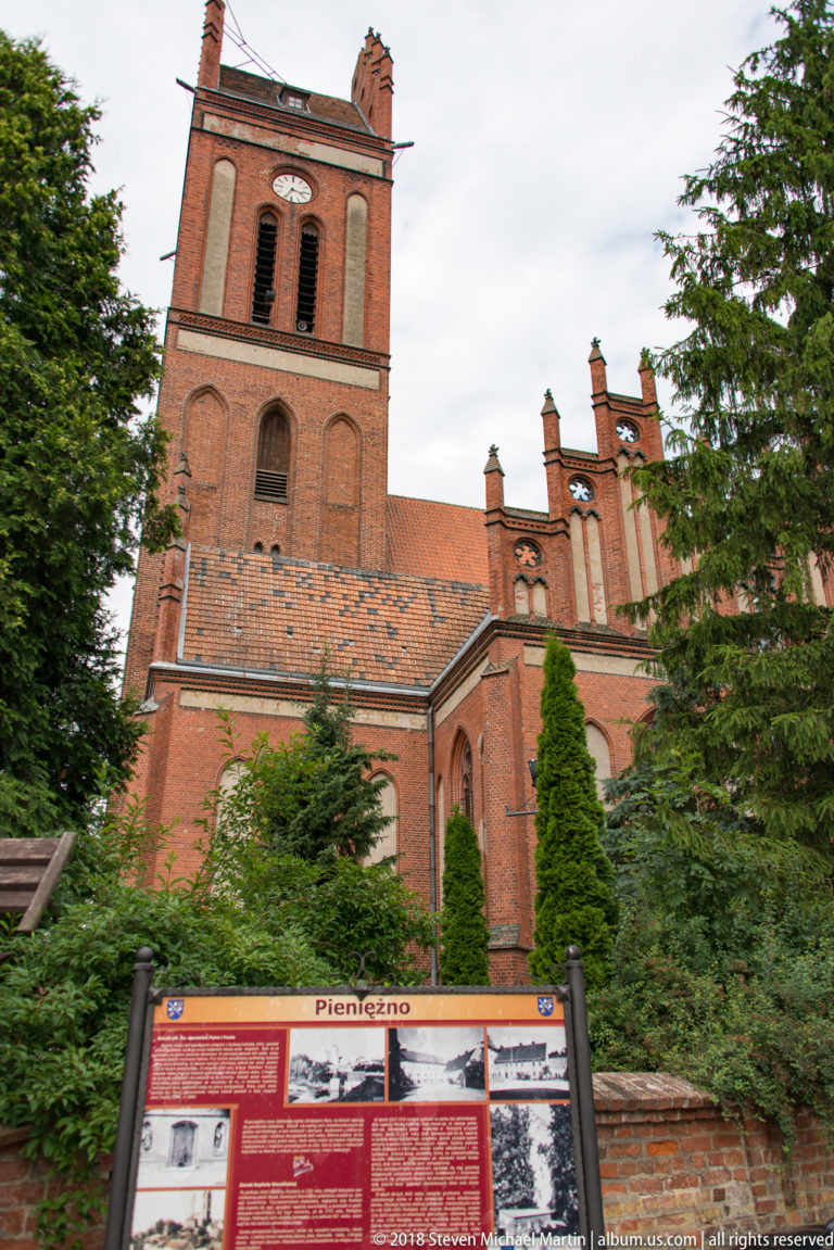 Red-brick Church of Saints Peter and Paul behind a foreground interpretive panel headed Pieniężno and landscaped shrubs.