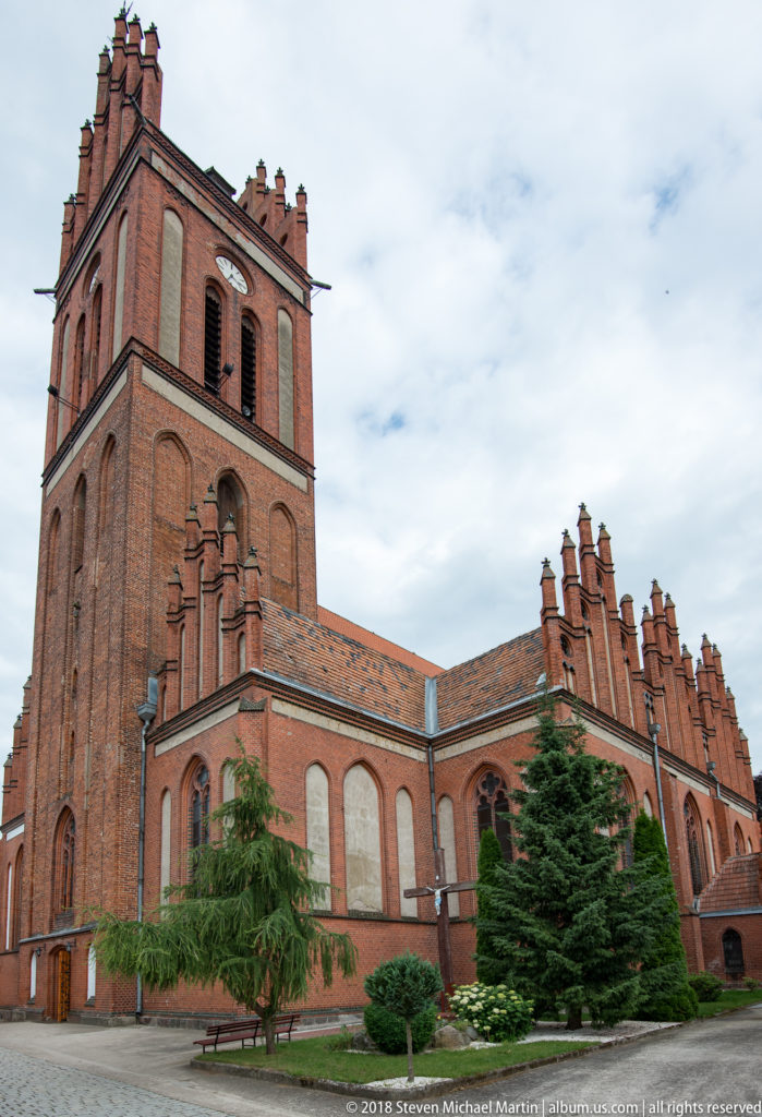 Side view of the red-brick Church of Saints Peter and Paul in Pieniężno, with square tower, pointed openings, and landscaped grounds.