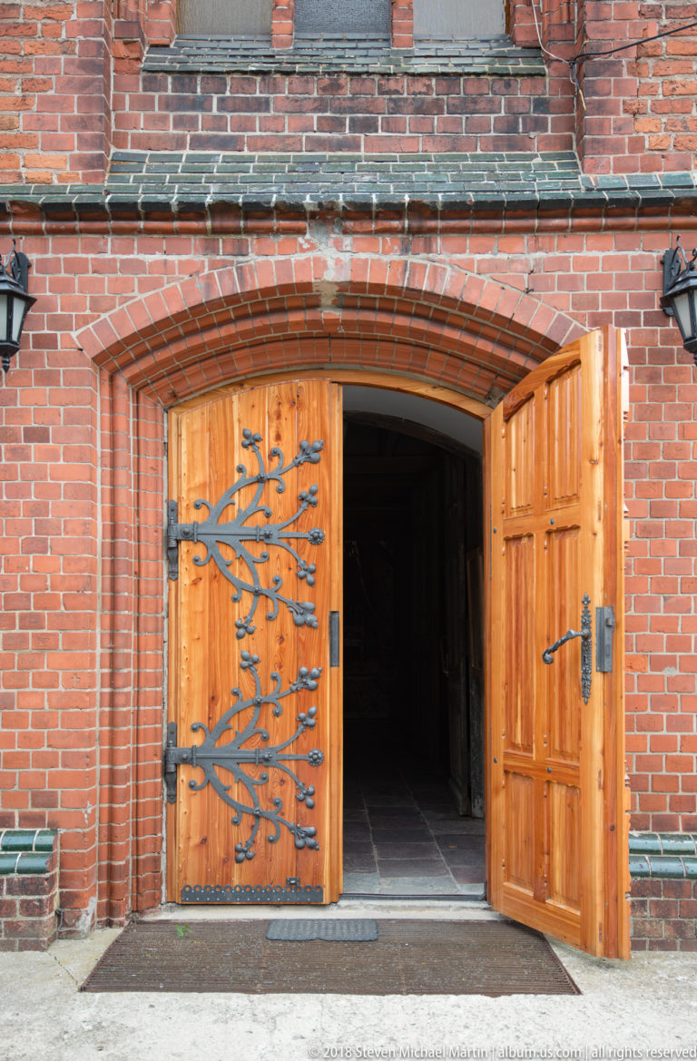 Open wooden double doors set in a broad red-brick arch, with black decorative strap hinges and a dark church interior beyond.