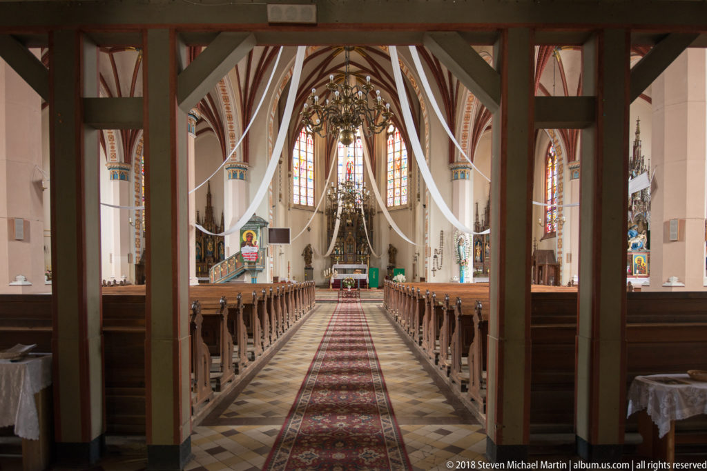 Central view down the nave of Saints Peter and Paul Church in Pieniężno, with pews, pointed vaults, stained glass, and white fabric swags.