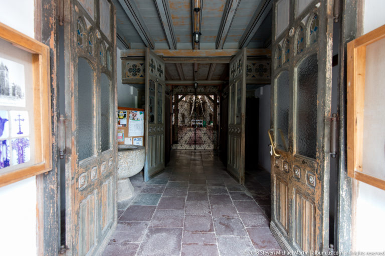 Open wooden church doors leading into a dim vestibule with decorative ironwork, glazed inner doors, stone floor, and holy-water basin.
