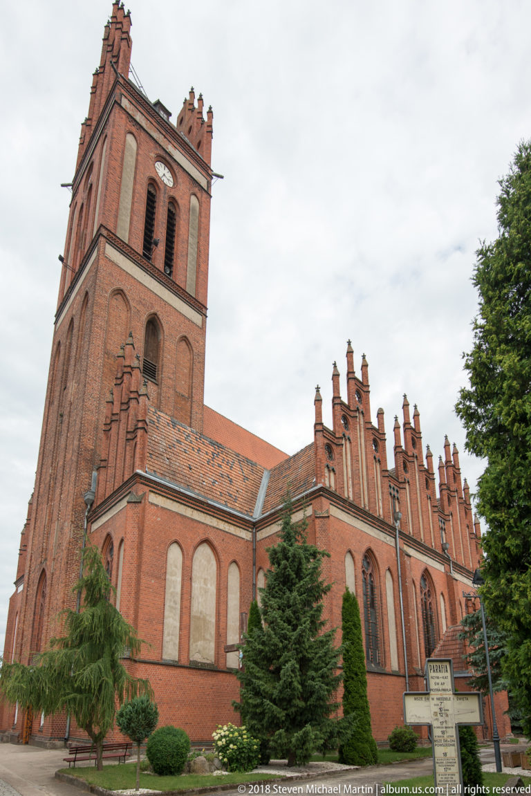 Oblique side view of the red-brick Church of Saints Peter and Paul in Pieniężno with west tower and stepped gables.