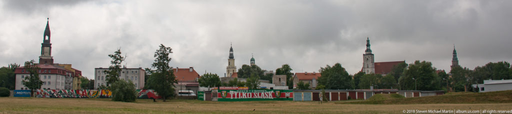 Distant Oleśnica skyline with several church and civic towers above red roofs, trees, fencing, and an open foreground under storm clouds.