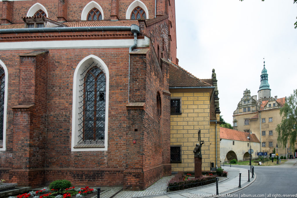 Brick Gothic church wall with pointed windows in the foreground and Oleśnica Castle tower visible beyond an arched passage.